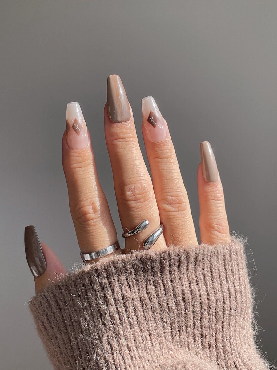 A hand with long coffin nails painted in glossy brown polish with two clear nails with brown and white lined diamond patterns
