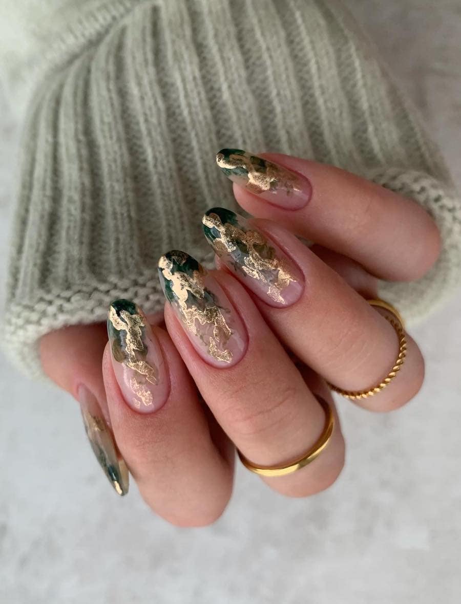 A hand with long almond nails painted with dark green and shimmering gold polish in a smoky pattern