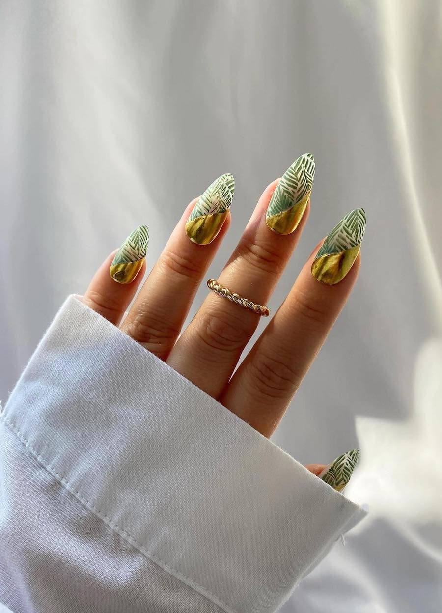 A hand with long almond nails with slanted gold polish and green floral nail art