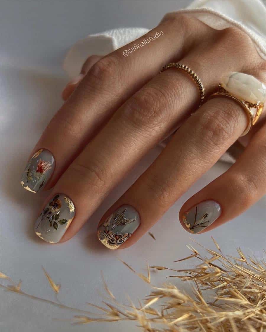 A hand with short square nails painted a sage green with floral and butterfly accents and gold flakes