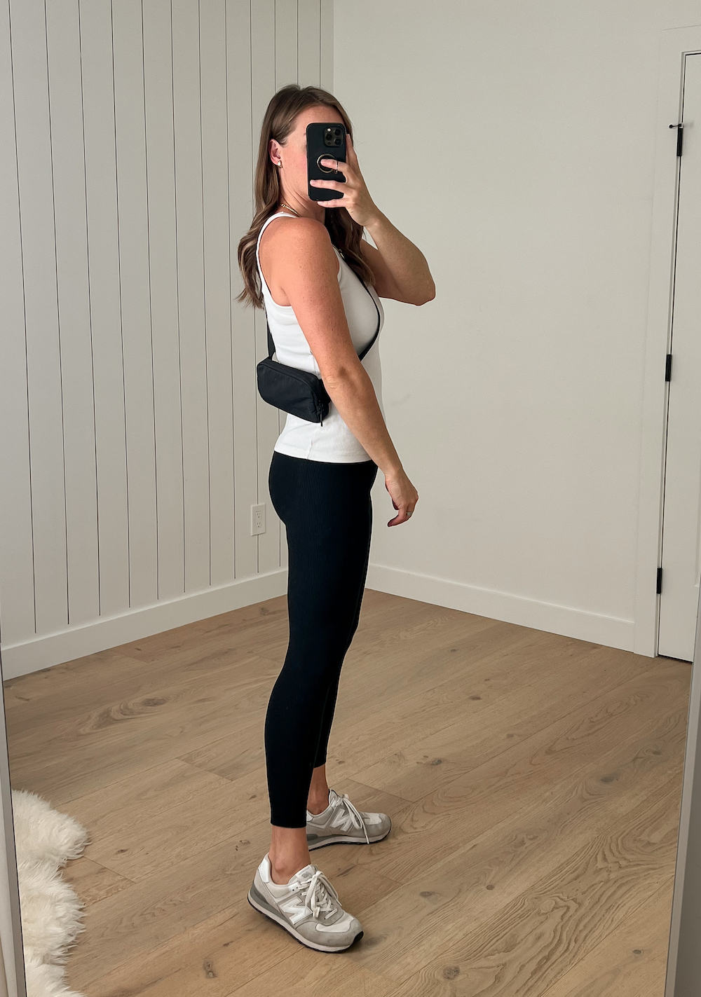 Christal wearing a white tank top, black leggings, white sneakers, and a black Lululemon belt bag crossbody style with the belt bag behind the back