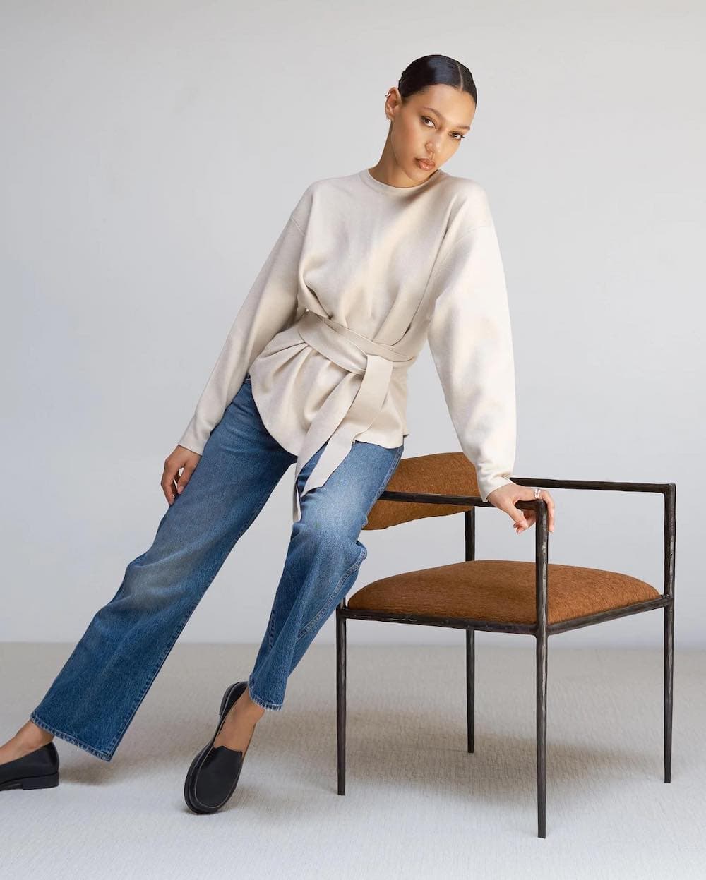 woman leaning on a leather chair wearing a cream sweater and blue jeans from Modern Citizen