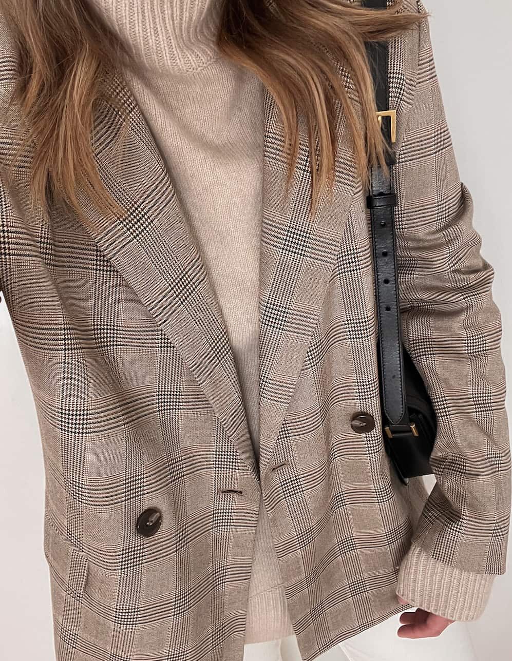 Up close shot of a woman wearing white jeans, a tan turtleneck and a tan and black plaid blazer.