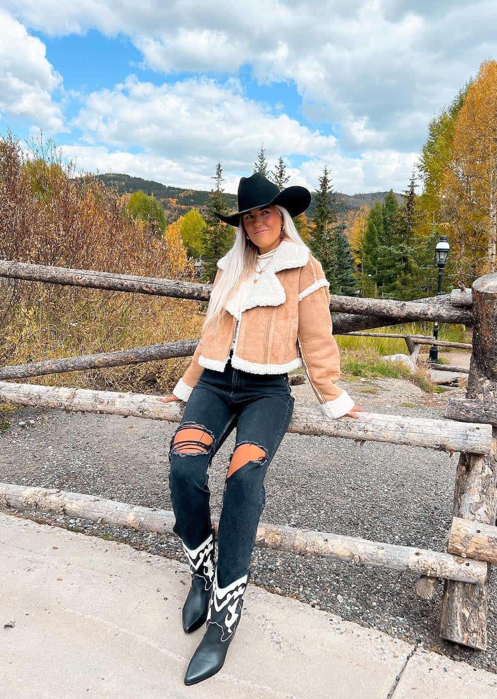 Woman wearing black ripped jeans with black and white cowboy boots, a suede and sherpa jacket and a black cowboy hat.