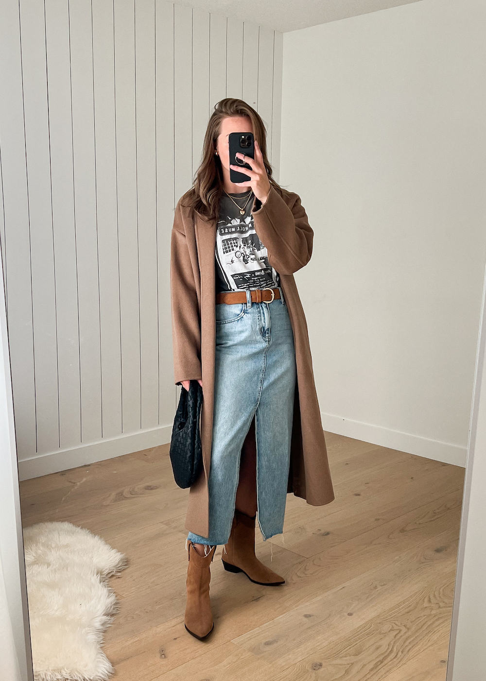 Woman wearing a denim maxi shirt, brown western style boots, a graphic tee and a brown trench coat.