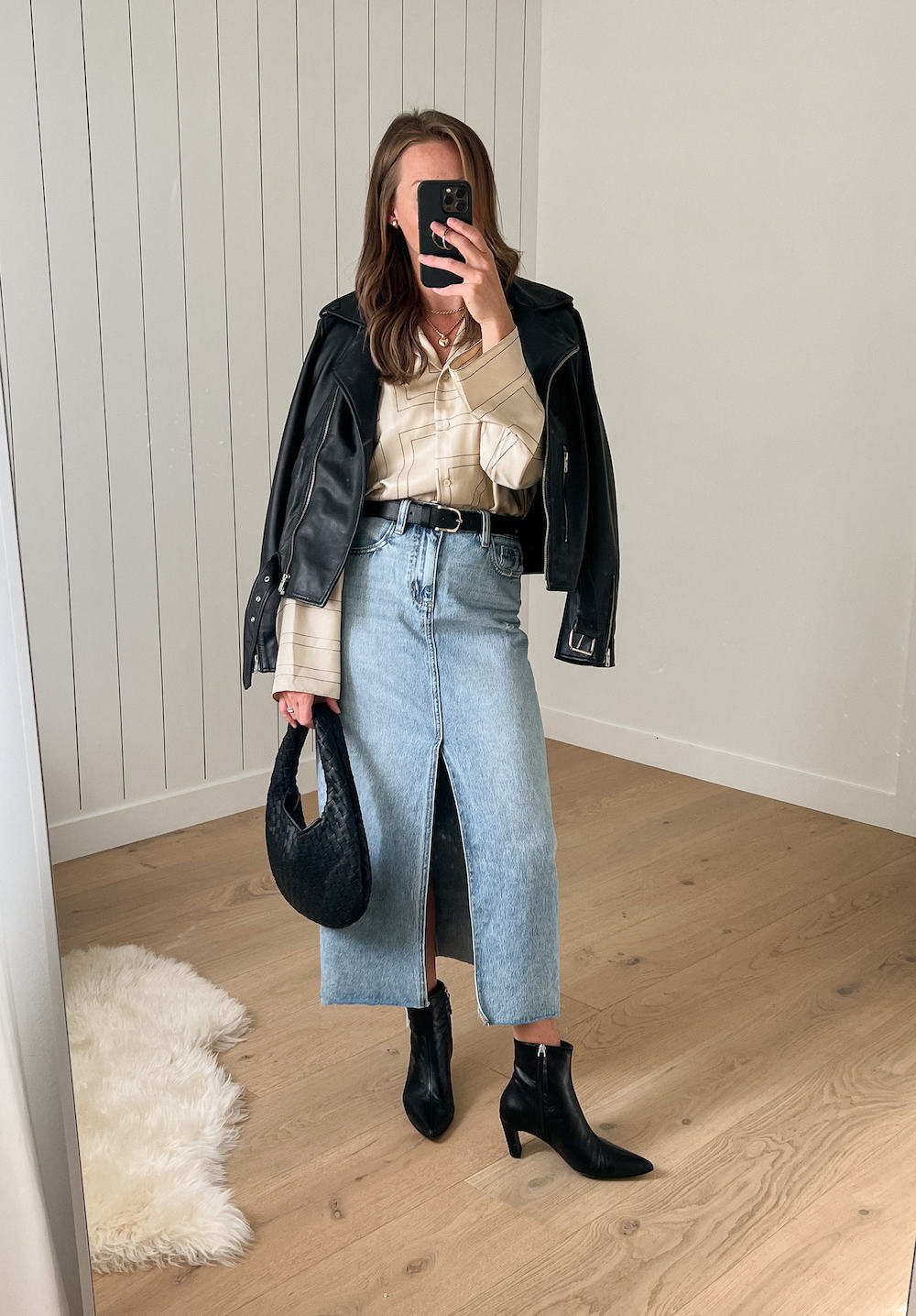 Woman wearing a denim midid skirt, black boots, cream button down shirt, and a black leather jacket.