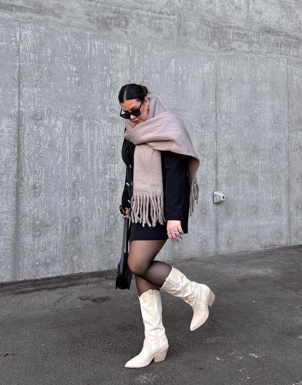 Woman wearing a black sweater dress with a large blanket scarf, black tights and white boots.