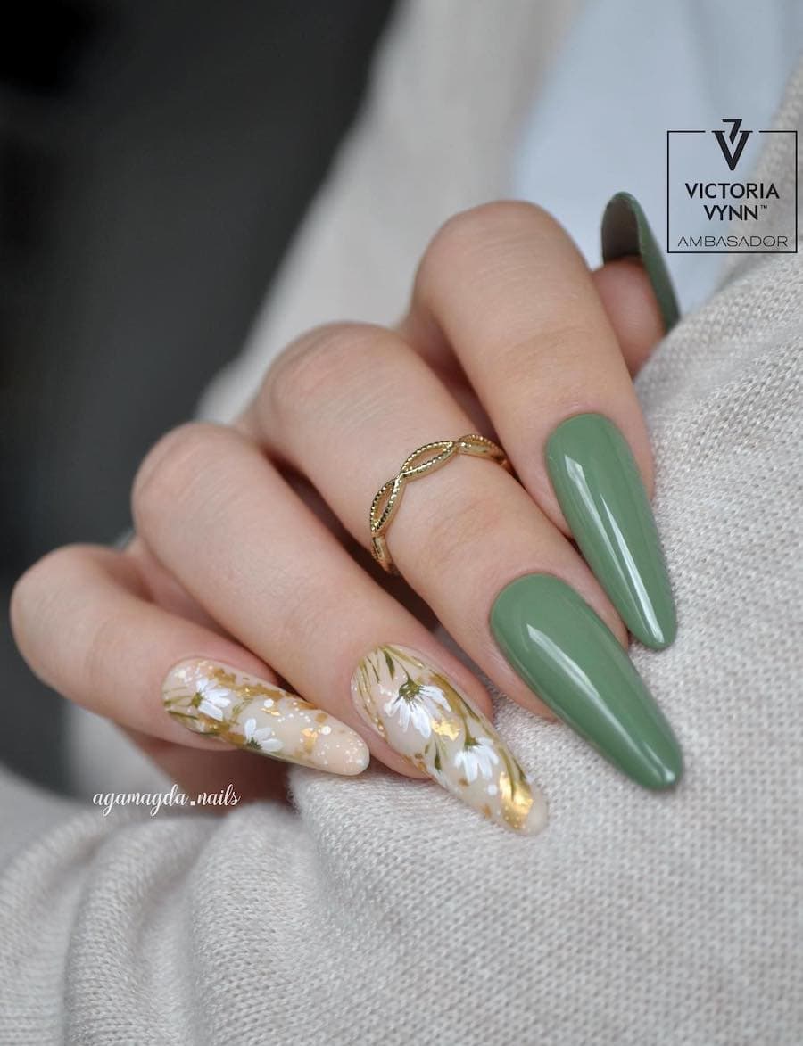 A hand with long almond nails painted solid green with two white and gold floral accent nails