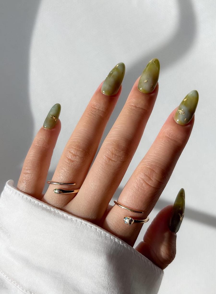 A hand with medium almond nails painted a marbled jade green with gold foil details