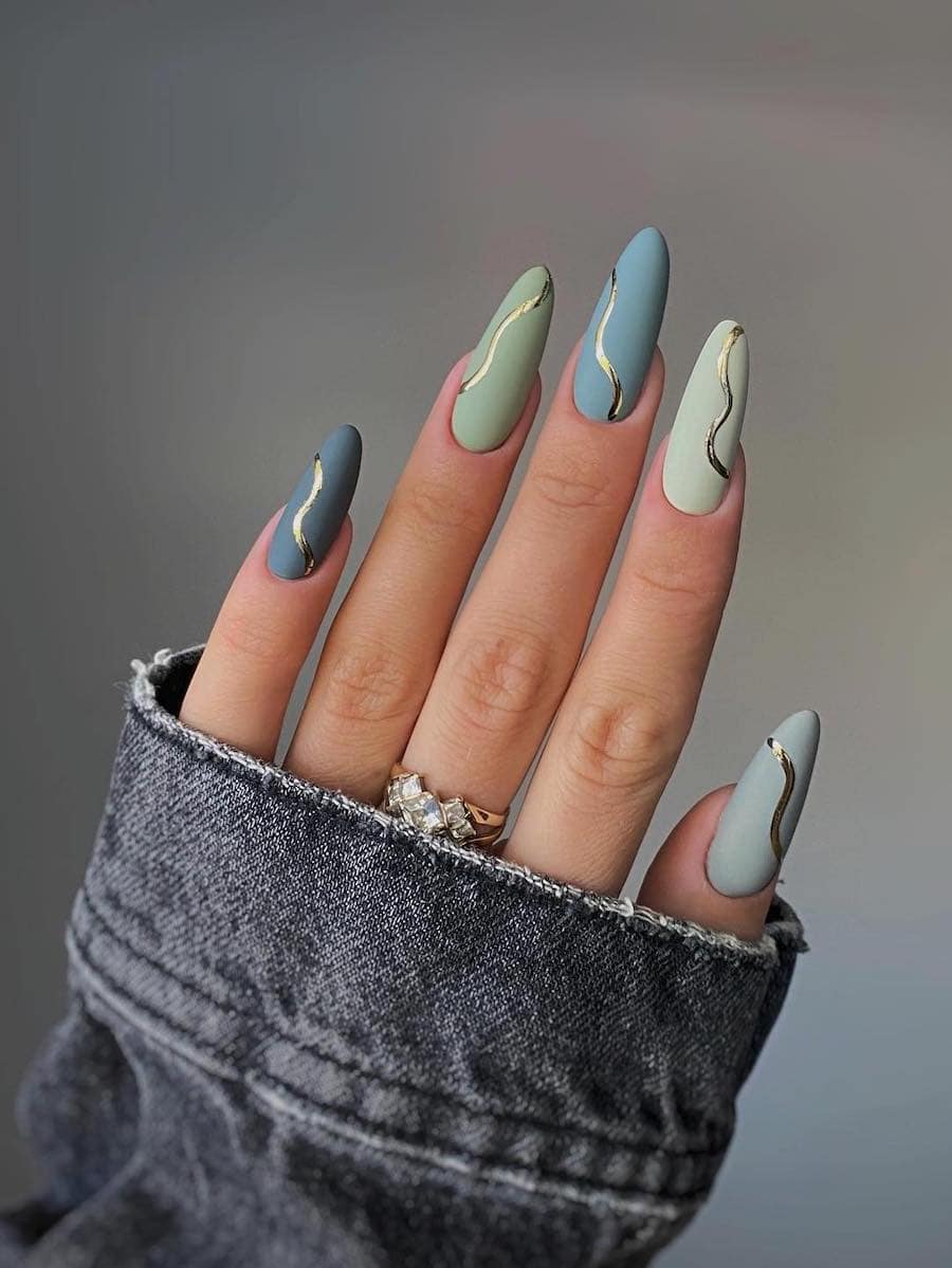 A hand with long almond nails painted in shades of green and blue with a matte finish and gold waves