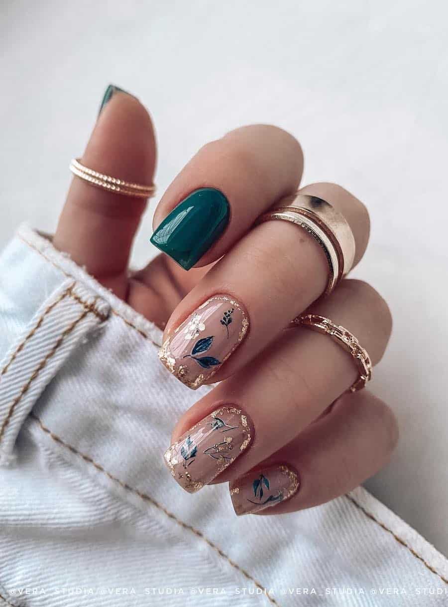 A hand with short square nails painted a glossy nude with green botanical art and gold foil borders and solid green accent nails