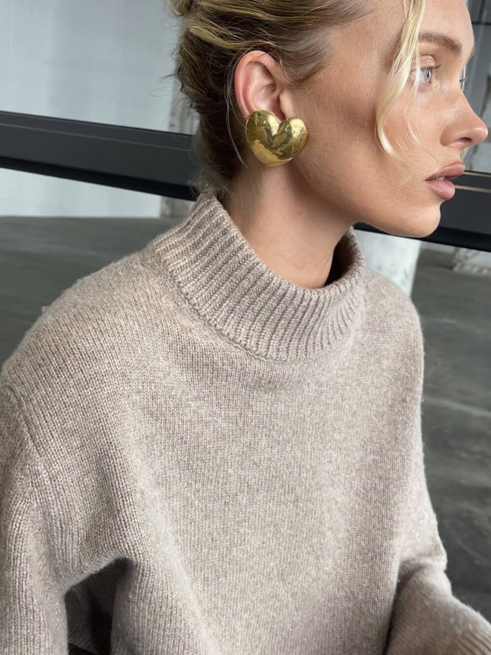 Woman wearing a chunky mock neck sweater with gold heart earrings and a fresh faced makeup look.
