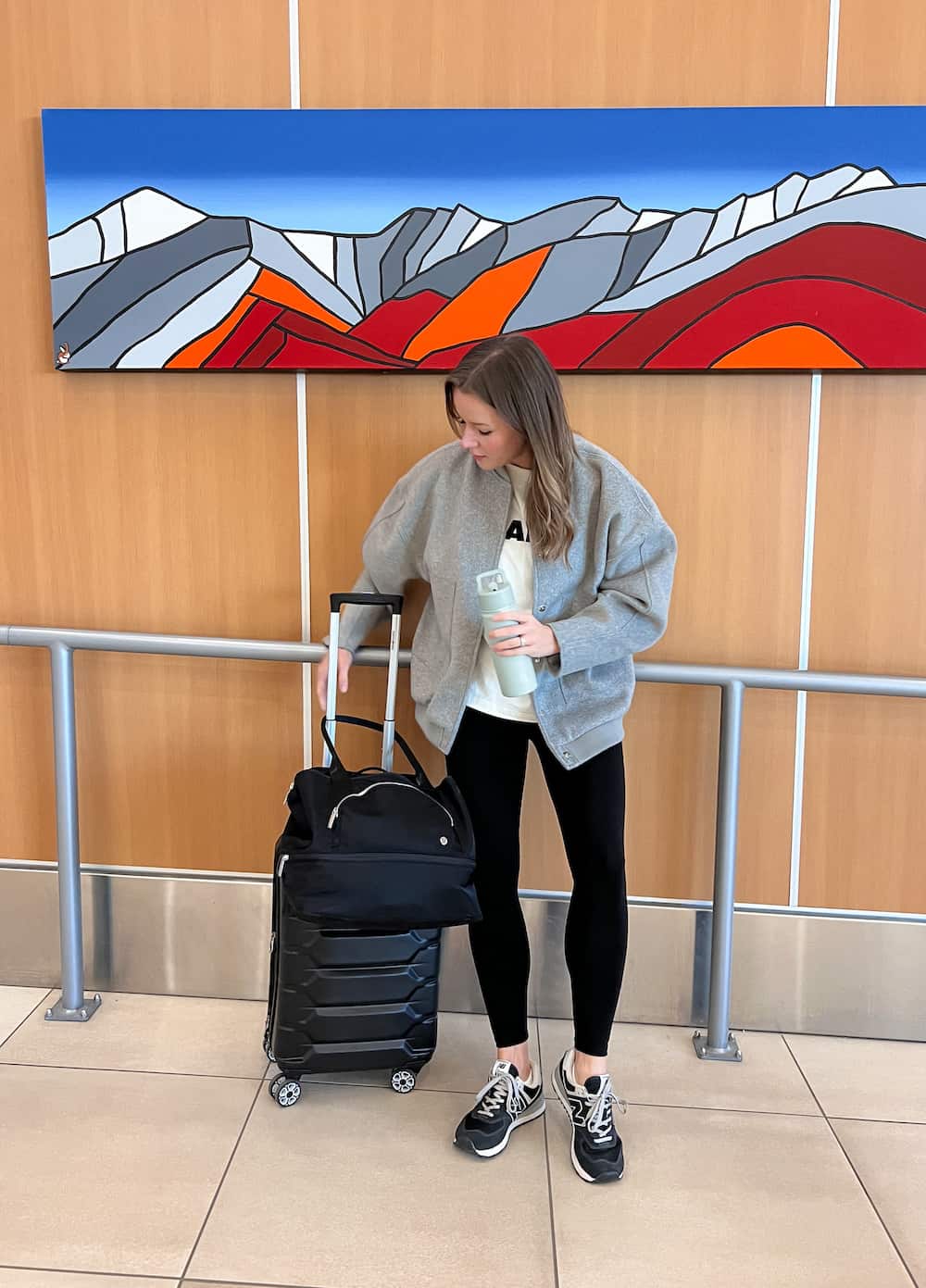 A woman at the airport wearing black leggings, a white graphic tee, a grey jacket, and black sneakers
