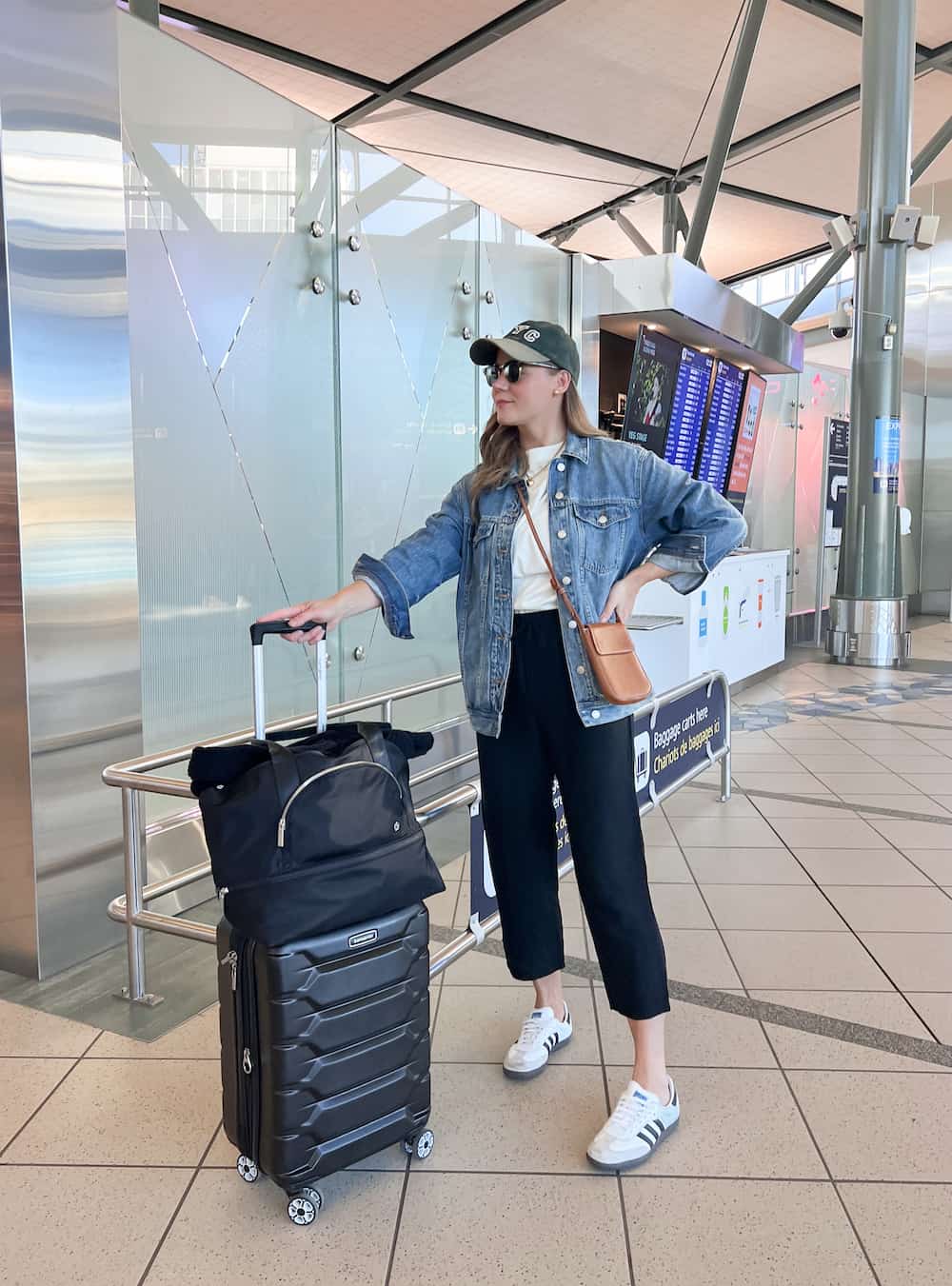A woman at the airport wearing black linen pants, a white tee, a denim jacket, white and grey sneakers, with a tan leather crossbody