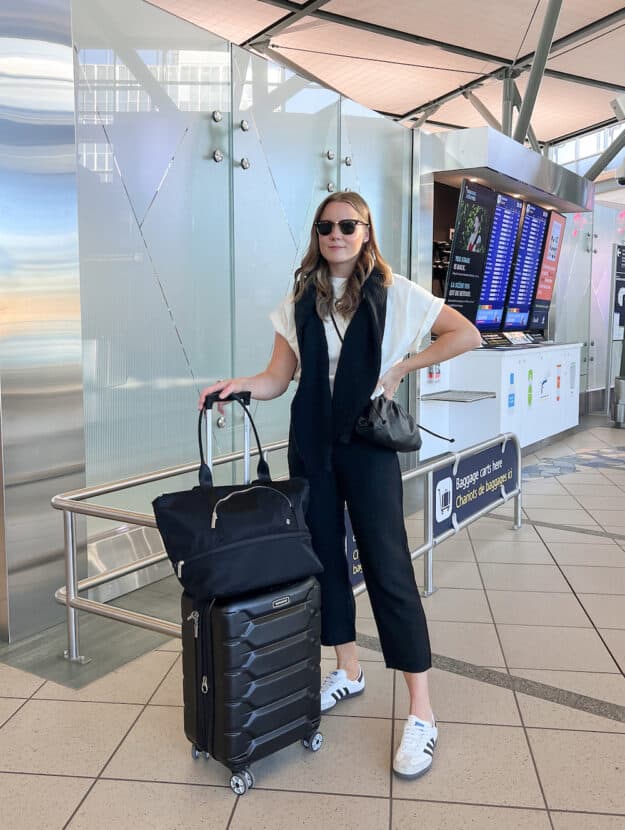 A woman at the airport wearing black linen pants and a white tee with a black sweater draped over her shoulders, grey and white sneakers, and a black crossbody bag