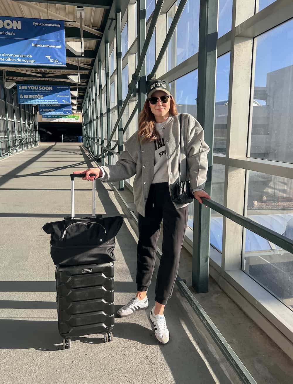 A woman at the airport wearing black joggers, a grey crewneck sweatshirt, a great jacket, white and grey sneakers, with a baseball cap and a black crossbody bag