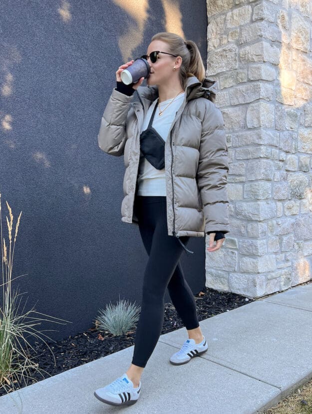 woman wearing an Aritzia Super Puff dupe jacket in taupe with a white top, black leggings and white sneakers