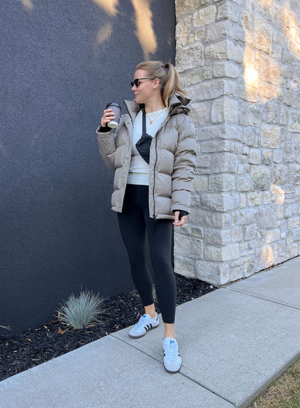 woman wearing a casual outfit with a taupe puffer jacket, white top, black leggings, and white Adidas Samba sneakers