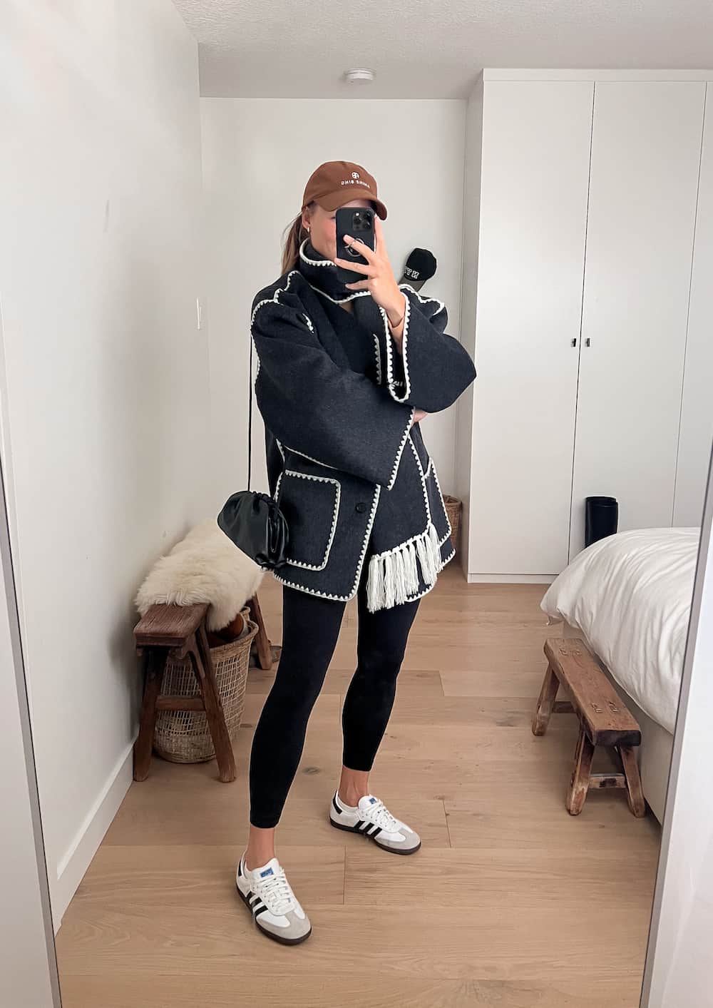 Woman wearing a dark grey oversized wool jacket with fringe and embroidery, black leggings, and white Adidas sneakers