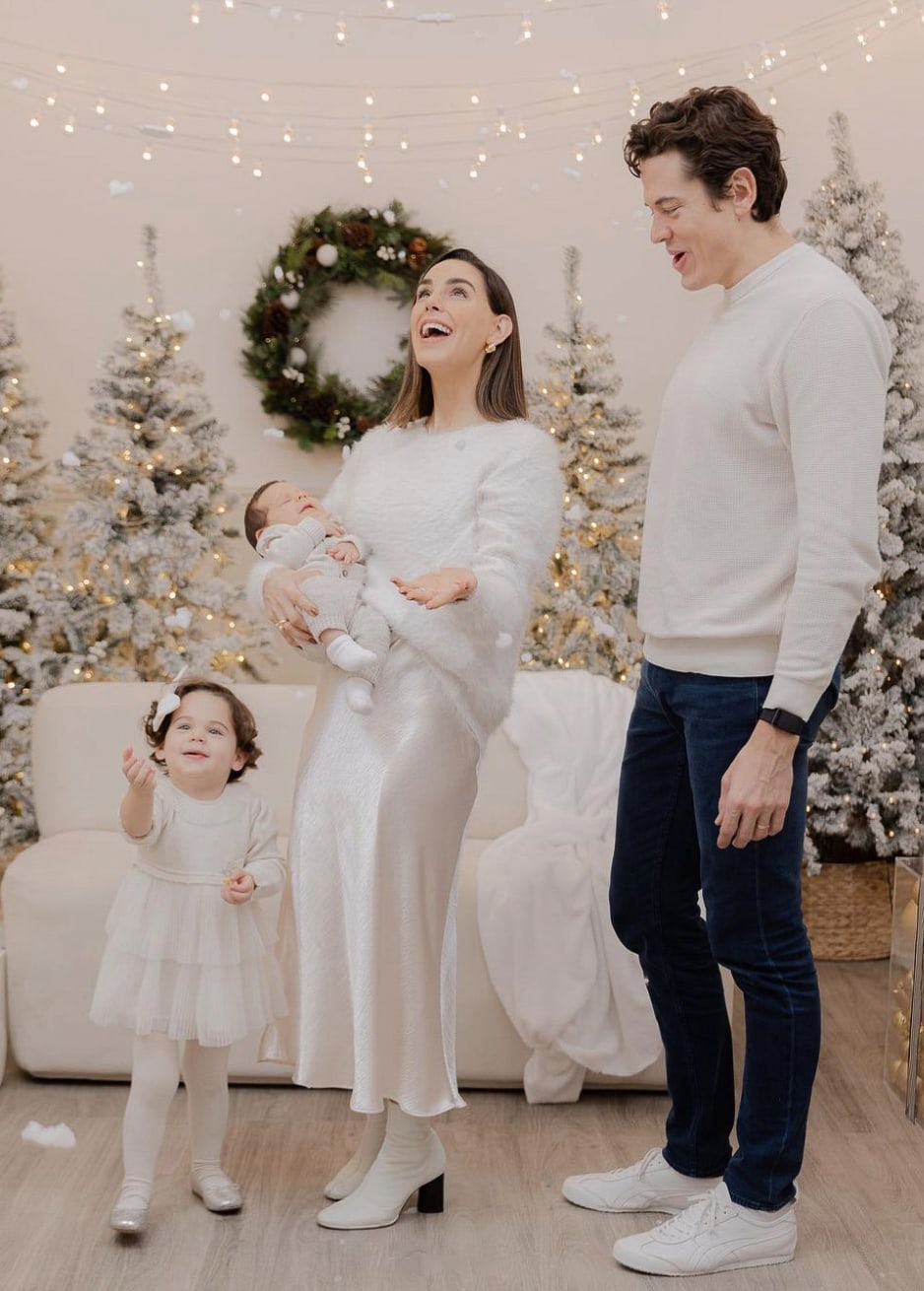 A Christmas family photoshoot with a family of four wearing all-white outfits with snowy Christmas trees in the background