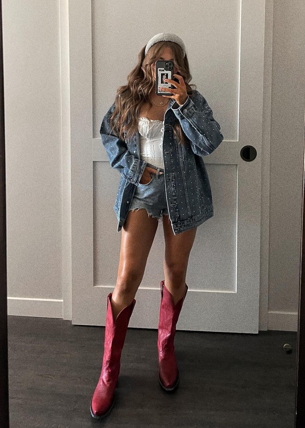 woman wearing a country concert outfit with an oversized jewelled denim jacket, white top, denim shorts, and red cowboy boots