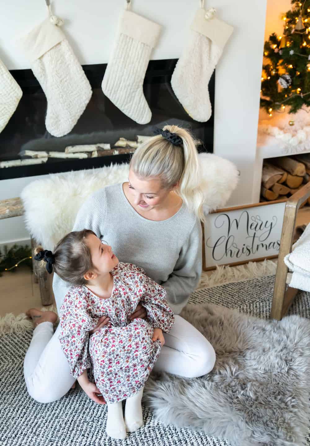 A Christmas family photo of a mom wearing white pants and a grey sweater and her daughter wearing a floral dress
