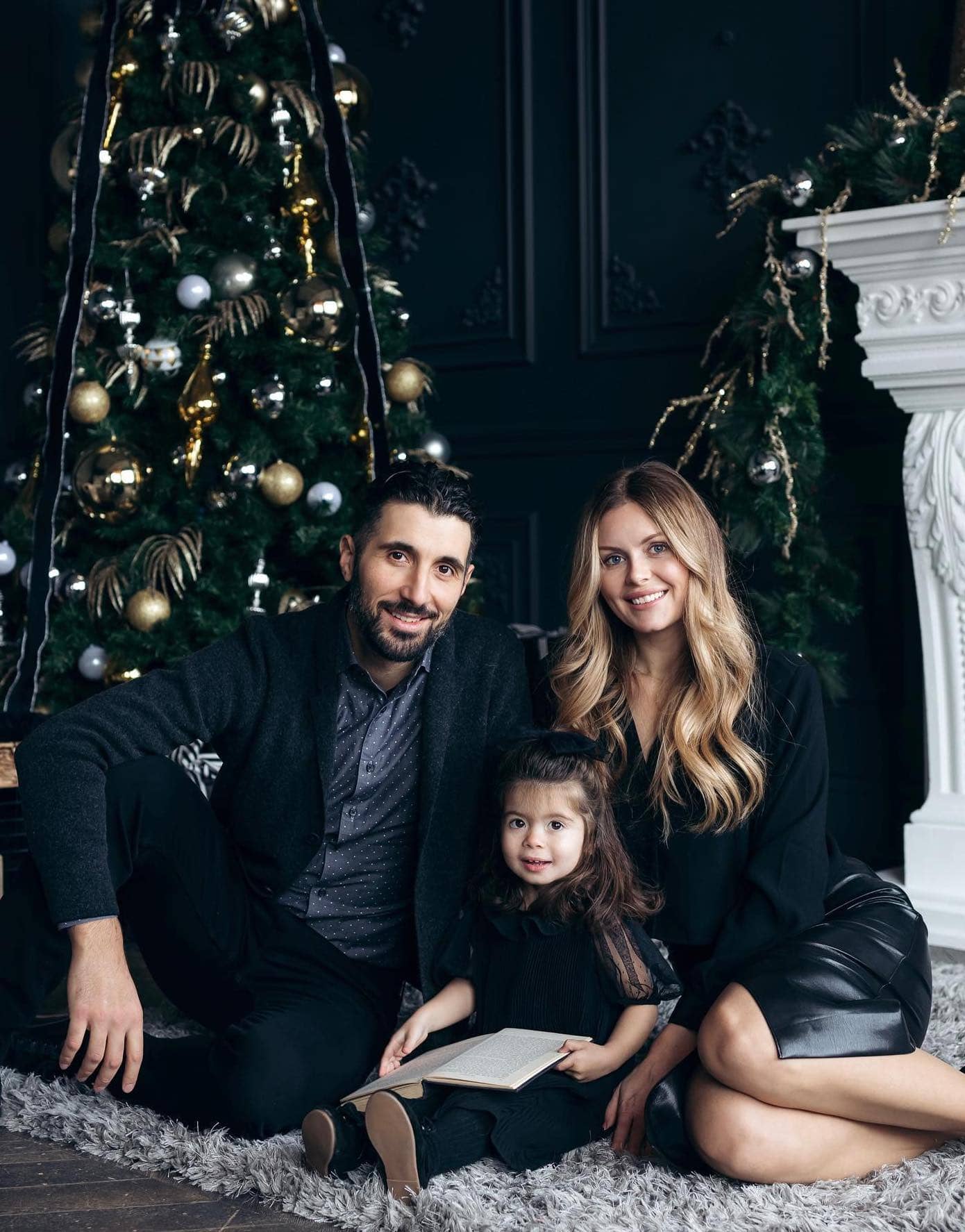 A Christmas family photo with a family of three sitting in front of a tree wearing dressy black pieces