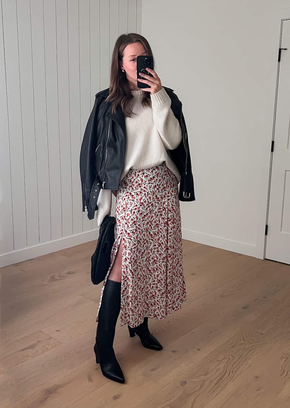 Christal wearing a floral printed dress with a white sweater over and black tall boots. She is also wearing a leather jacket.