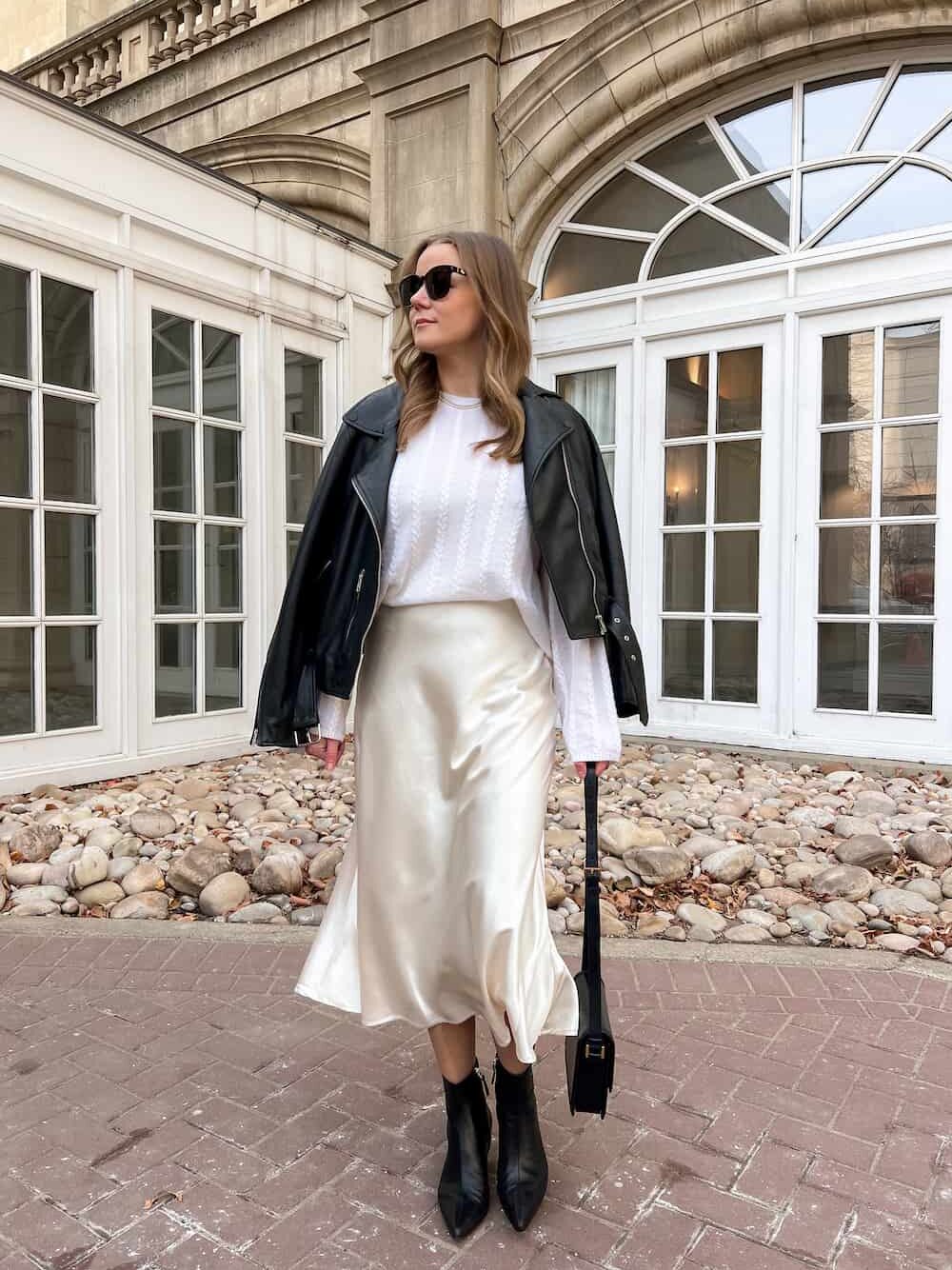 Christal wearing a holiday outfit with a leather jacket, Lilysilk ivory cable knit sweater, ivory slip skirt, and black leather ankle boots