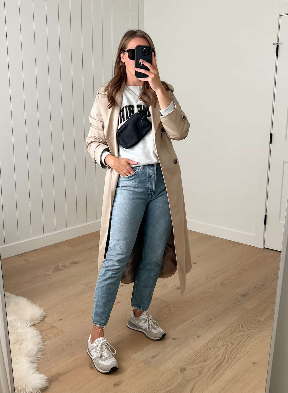 woman wearing a long tan trench coat over a grey sweatshirt and jeans with white New Balance sneakers 