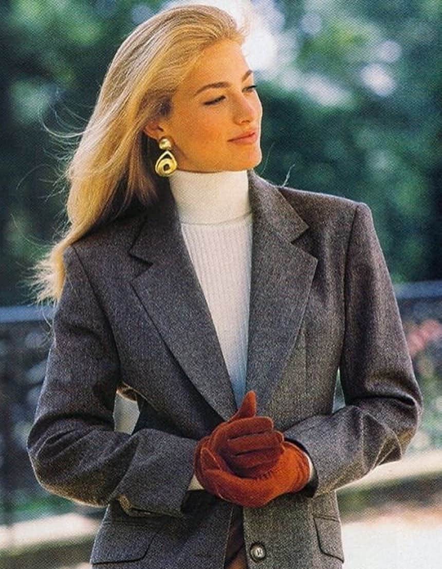 A woman with an old money aesthetic wearing a grey blazer over a fitted white turtleneck with red gloves and gold earrings