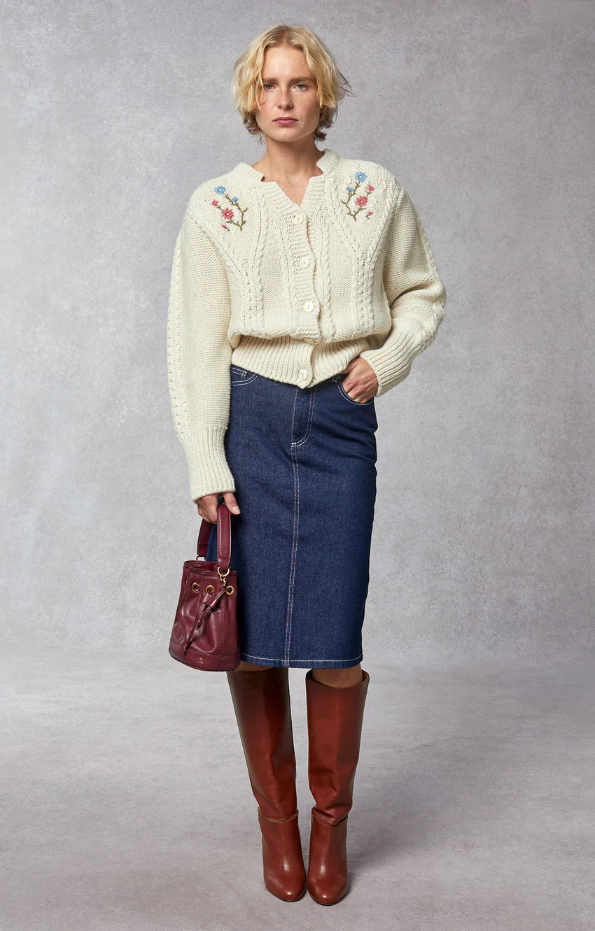 Woman wearing a denim midi skirt with tall brown boots and a white embroidered cardigan.
