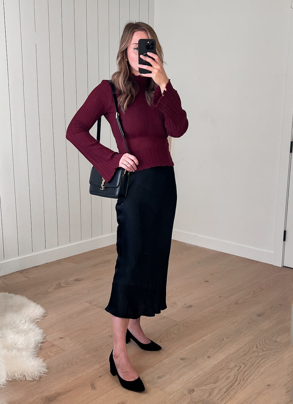 A woman wearing a black silk midi skirt with a red knit crewneck sweater, black heels, and a black shoulder bag