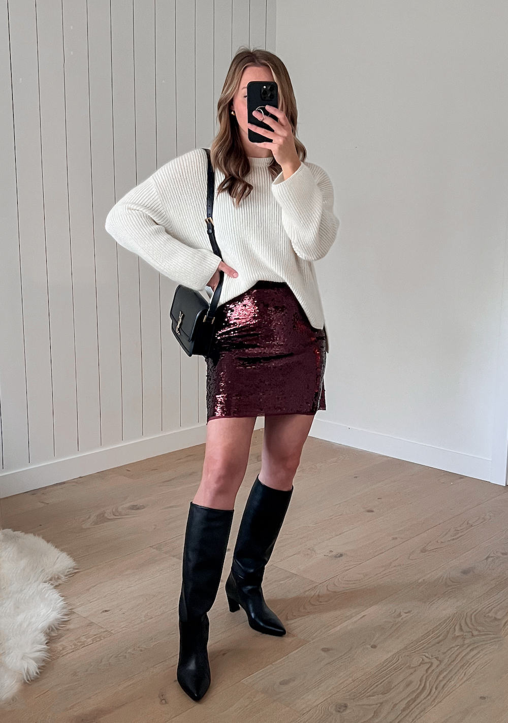 A woman wearing a sparkly burgundy mini skirt with a white knit crewneck sweater, a black shoulder bag, and knee-high black leather boots