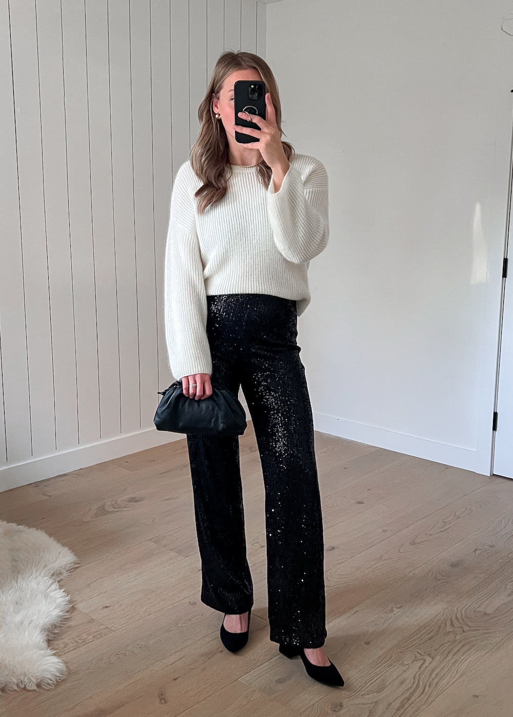 A woman wearing sparkly black wide-leg pants with a white knit crewneck sweater, black heels, and a dark green clutch