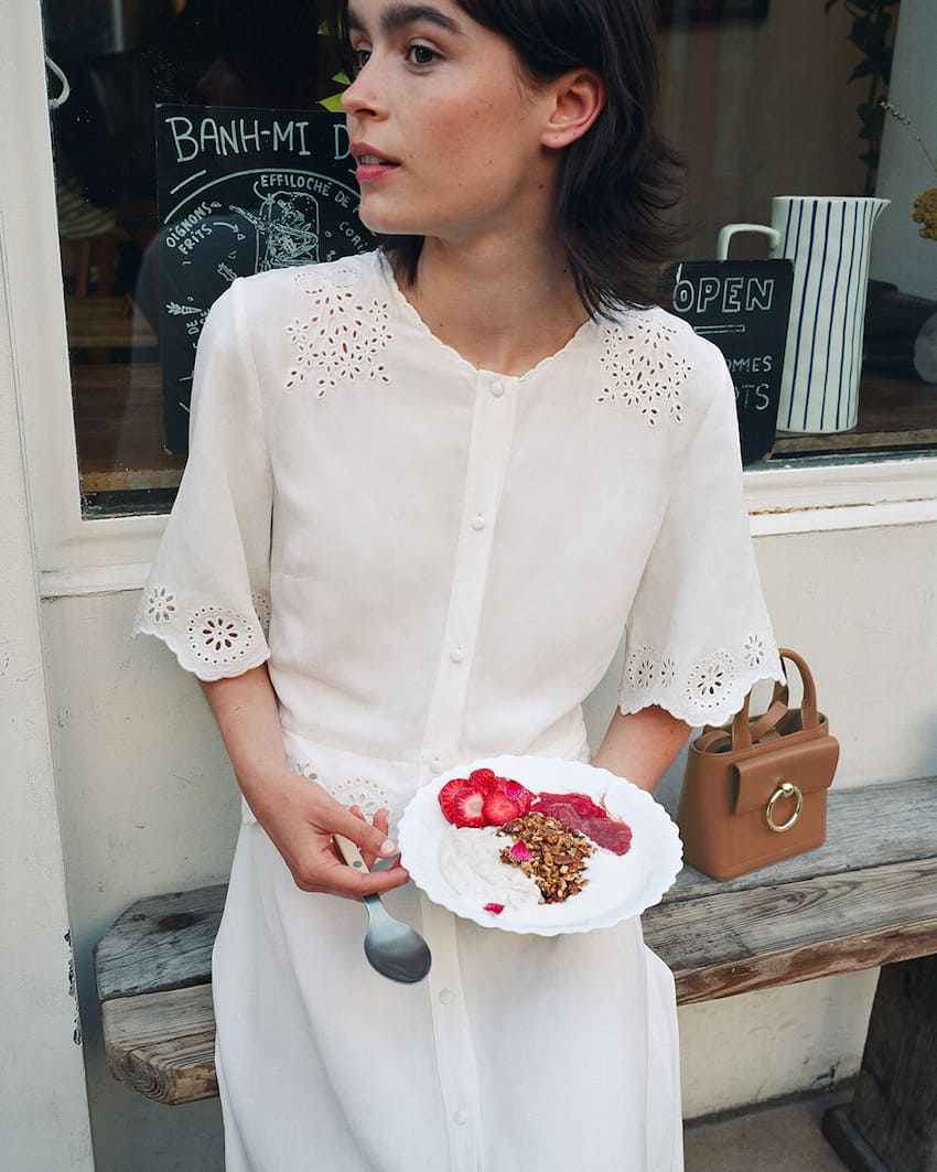 Woman wearing a white eyelet dress while eating breakfast outside of a cafe.