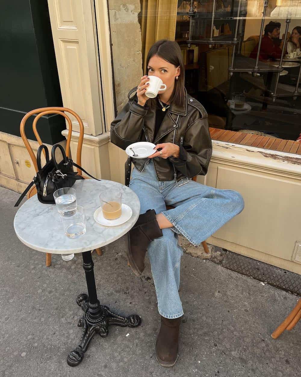 French woman wearing an outfit in Paris at a cafe with a leather jacket, blue jeans, and dark brown Ugg boots