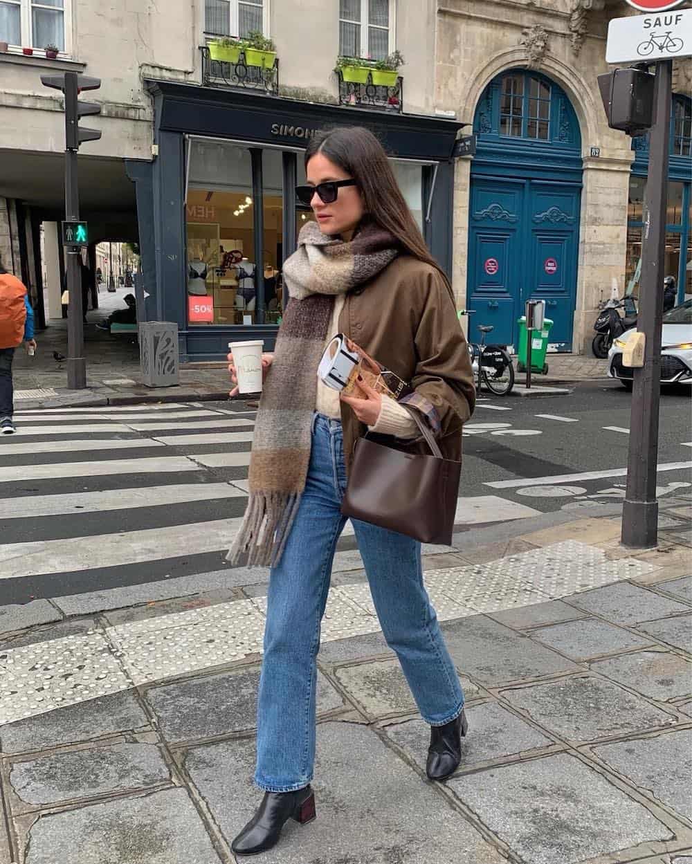 French woman wearing an outfit with a brown jacket, checkered neutral scarf, blue jeans, and brown ankle boots in Paris