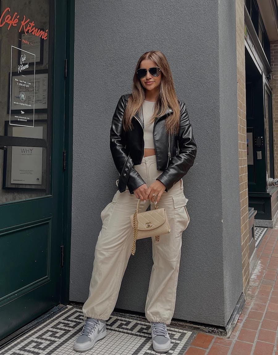 Woman wearing off white cargo pants with a white cropped top and a black leather jacket and sneakers.