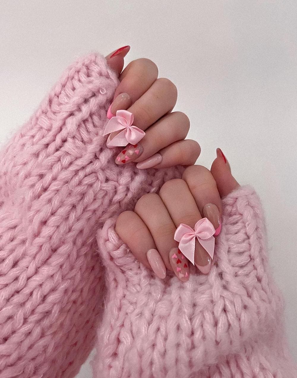 A hand with medium almond nails featuring pastel pink nails, pink French tips, heart accented nails, and satin bows