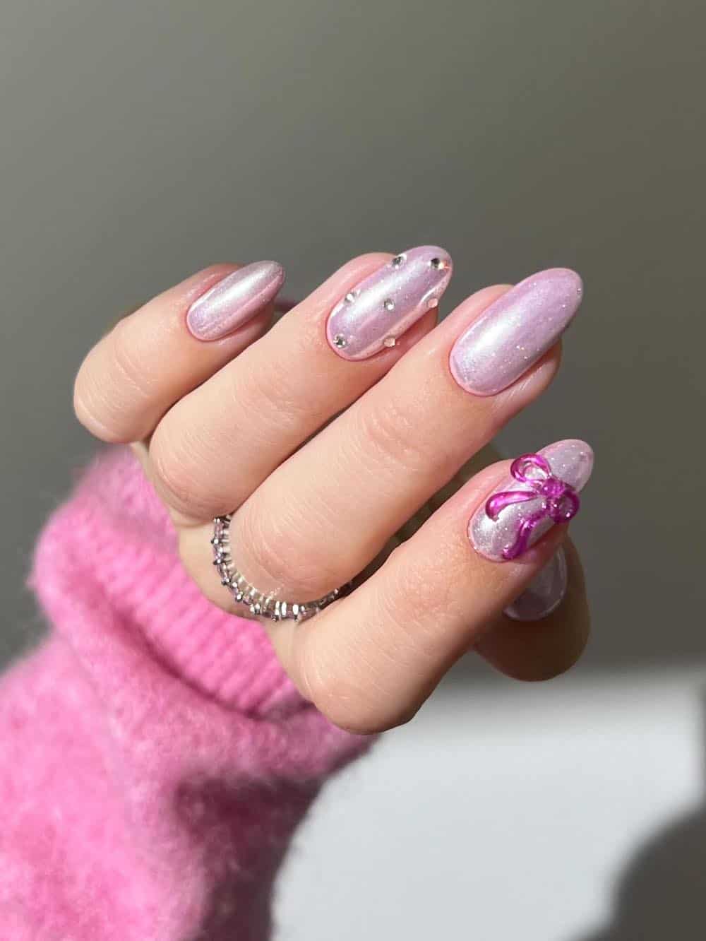 A hand with short almond nails painted a shimmering pink with gem accents and bright pink bow details