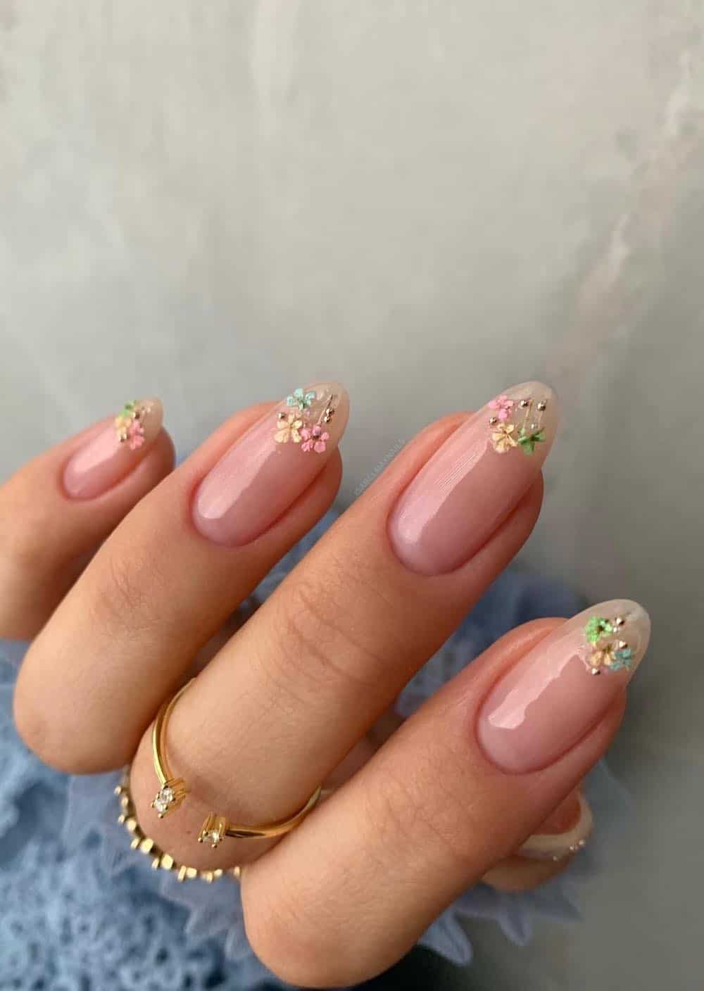 A hand with short glossy, nude almond nails with dainty pastel flowers and gold dot accents