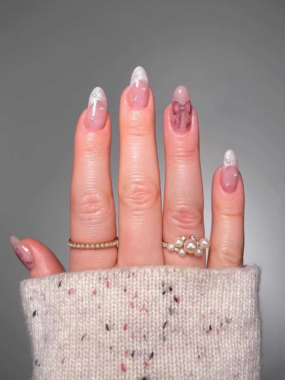 A hand with short almond nails with pearly tips and pearl accents and two light pink accent nails with bow nail art