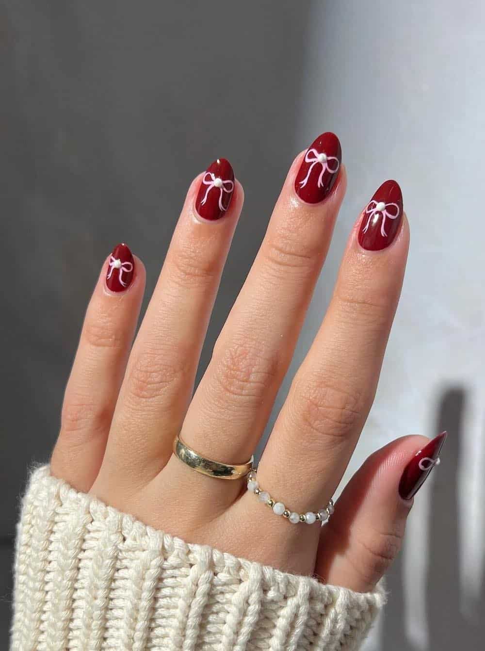 A hand with short almond nails painted a dark red with pink bows and pearl details