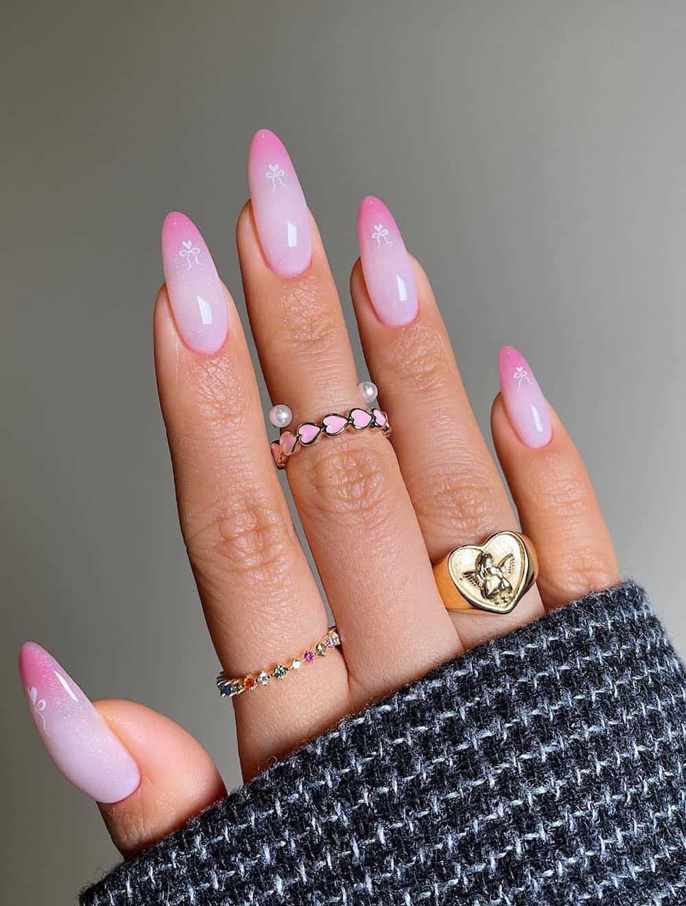 A hand with long almond nails painted a shimmering white with blended pink tips, white bow art, and white heart accents