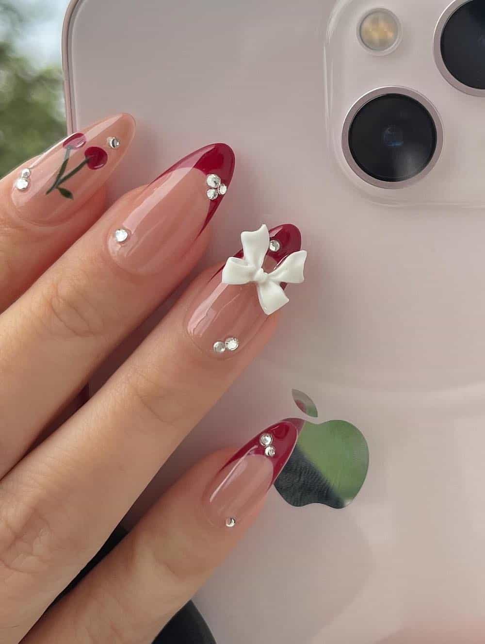 A hand with medium nude almond nails with red tips, cherry nail art, crystals accents, and white bow charms