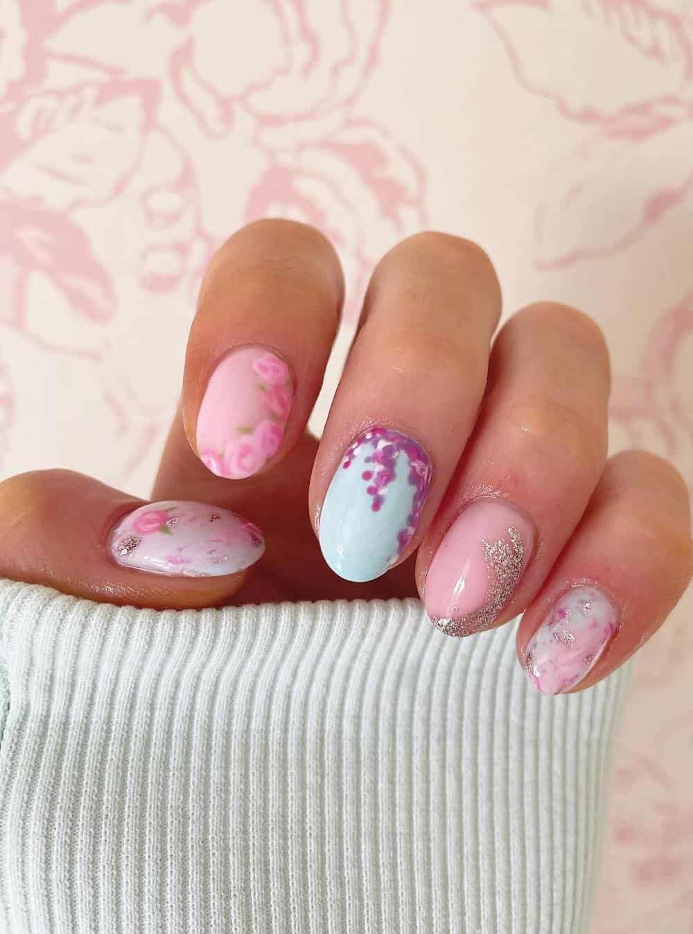 A hand with short round nails featuring pink, purple, and blue floral designs and glitter details