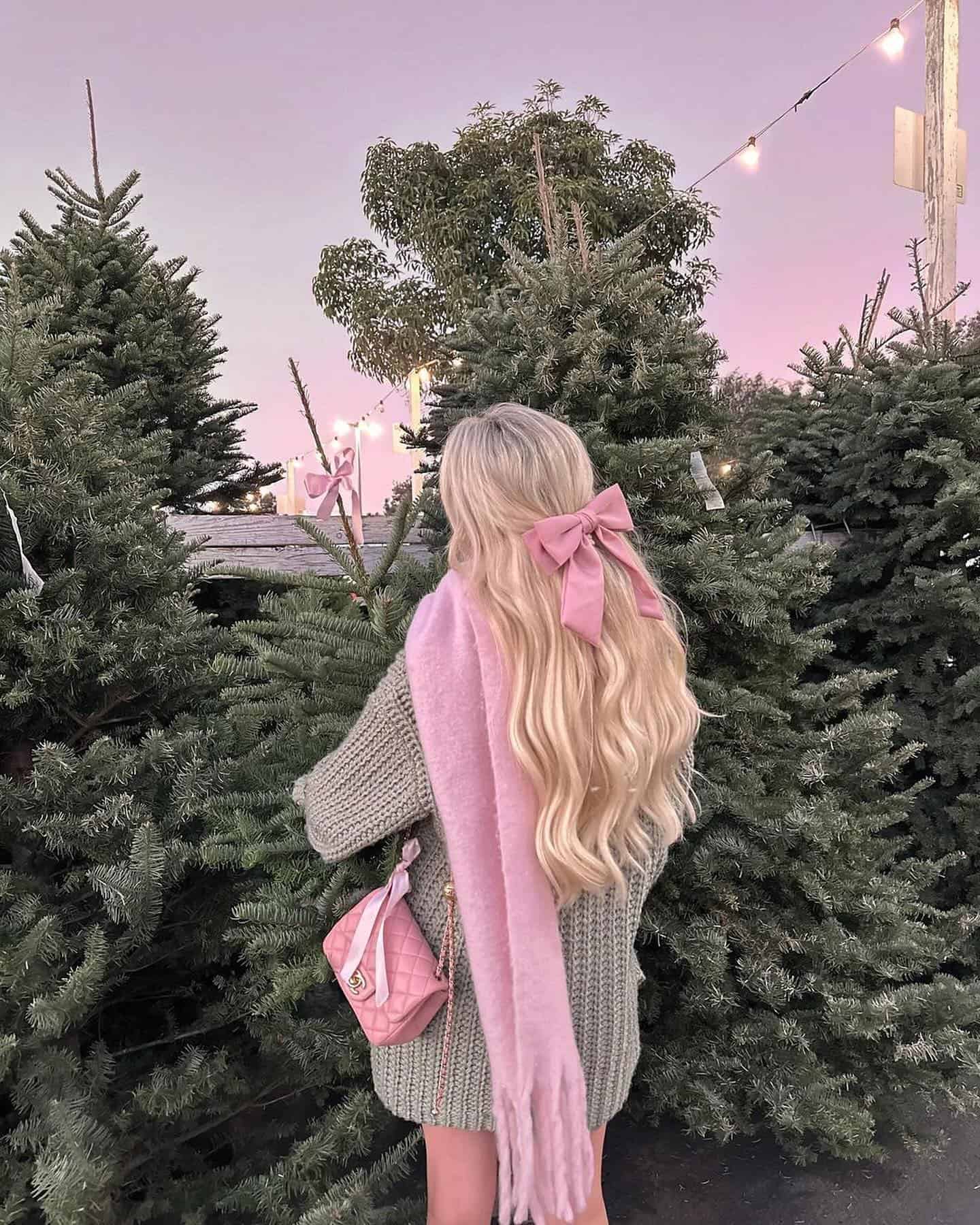 A woman wearing a sage green oversized sweater dress with a pink scarf, a pink hair bow, and a quilted pink crossbody purse
