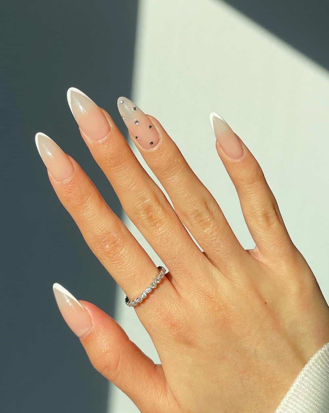 A hand with long almond nails painted with pointed white tips and a crystal accent nail