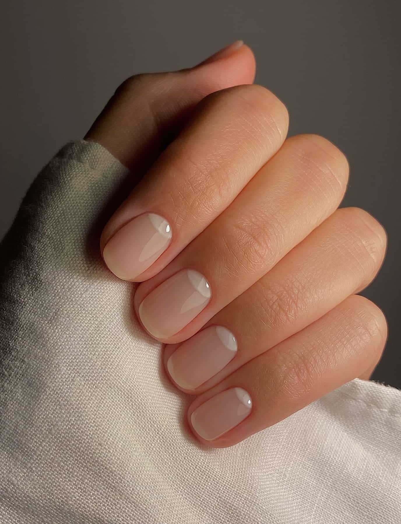 A hand with short squoval nails painted a glossy nude with white half moon details